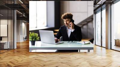 Young man working on laptop and talking on the mobile phone while sitting at his working place in office Wall mural